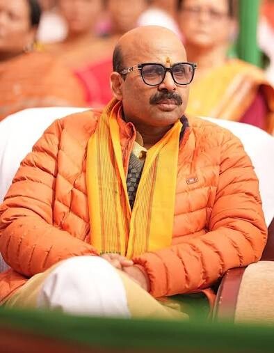 Harish Dwivedi seated at an outdoor public event wearing a winter jacket and a ceremonial stole