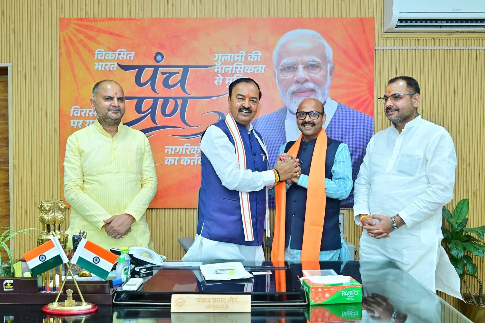 Harish Dwivedi at his office desk with colleagues, national flags and a party backdrop behind